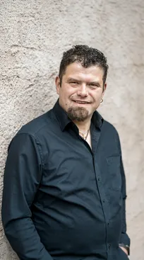 Michael Muggi Reiterer mit Kinnbart vor rustikaler Steinwand, goldene Halskette, lässig angelehnt