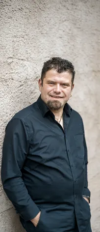 Michael Muggi Reiterer mit Kinnbart vor rustikaler Steinwand, goldene Halskette, lässig angelehnt