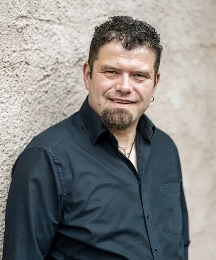 Michael Muggi Reiterer mit dunklen Locken und Kinnbart in schwarzem Hemd, goldene Halskette und Ohrring, herzlich lächelnd