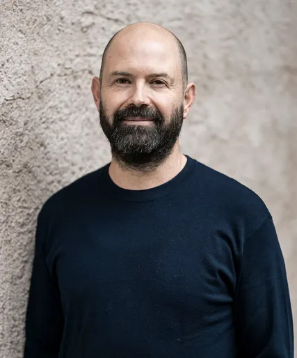 Wolfgang Tratter mit Glatze und Vollbart in blauem Pullover vor grau-beiger Steinwand, freundlich lächelnd, Teamportrait