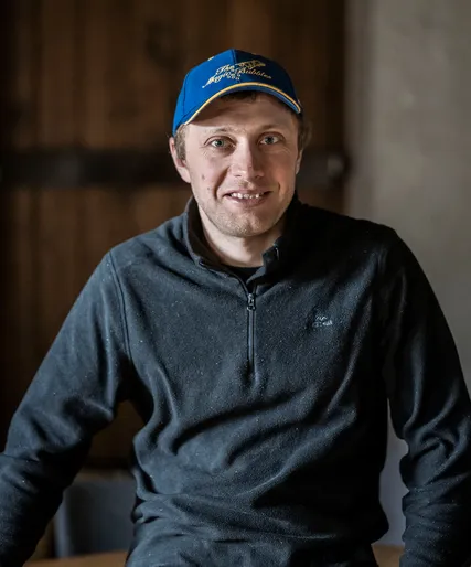 Mitarbeiter mit blauer Baseballkappe und Fleece-Pullover, freundlich lächelnd, Arunda-Teamportrait vor Holzwand