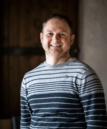 Mitarbeiter mit Dreitagebart in grau-blau gestreiftem Strickpullover, herzlich lächelnd, Arunda-Teamportrait vor Holzwand