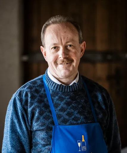 Senior-Mitarbeiter mit grauem Schnurrbart in blauer Arunda-Schürze, warmherzig lächelnd, Teamportrait vor Holzwand
