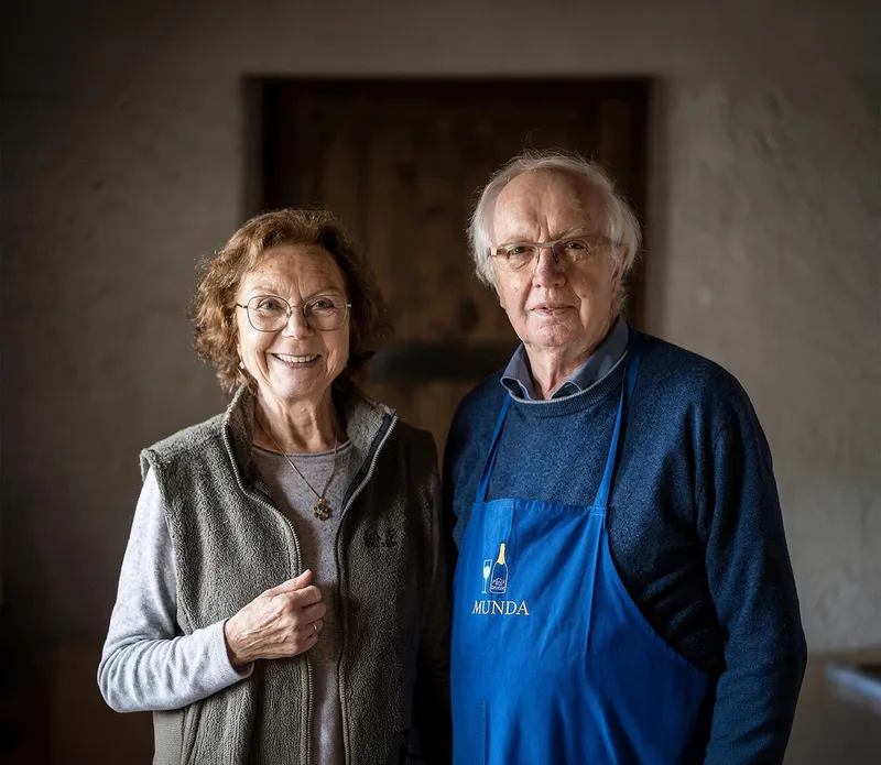 Gründerpaar Sepp und Marianne Reiterer in Arunda-Schürze vor grau-brauner Kellerwand, freundlich lächelnd, Mölten