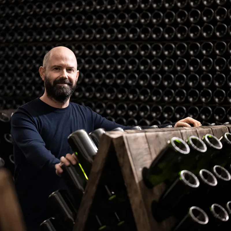 Lächelnder Mitarbeiter am Holz-Rüttelpult vor raumhoher Wand aus hunderten lagernden Sektflaschen im Arunda-Keller