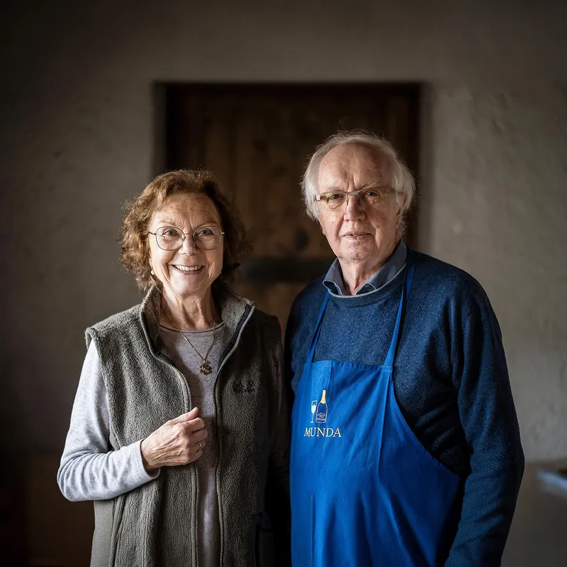 Gründerpaar Sepp und Marianne Reiterer in Arunda-Schürze vor grau-brauner Kellerwand, freundlich lächelnd, Mölten