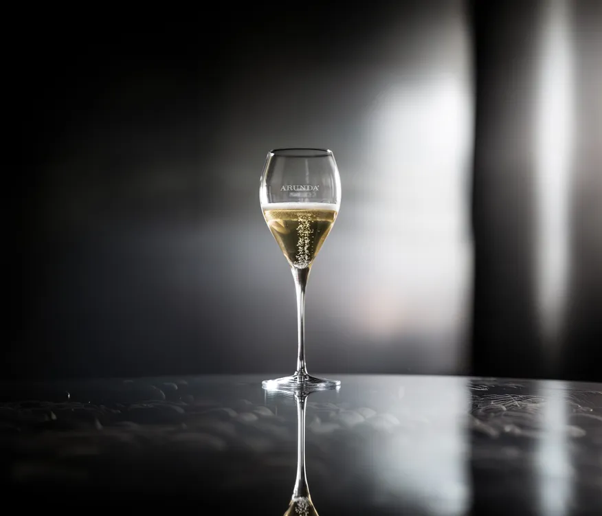 Tulpenförmiges Arunda-Sektglas mit goldenem Sekt und Perlage auf dunkler Spiegelfläche, dramatisches Spotlight