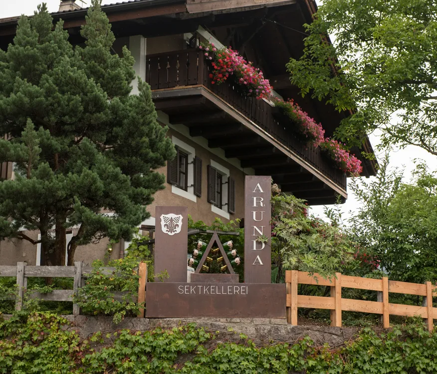 Traditionelles Südtiroler Haus mit Holzbalkonen und rosa Geranien, Arunda Sektkellerei Schild, Mölten
