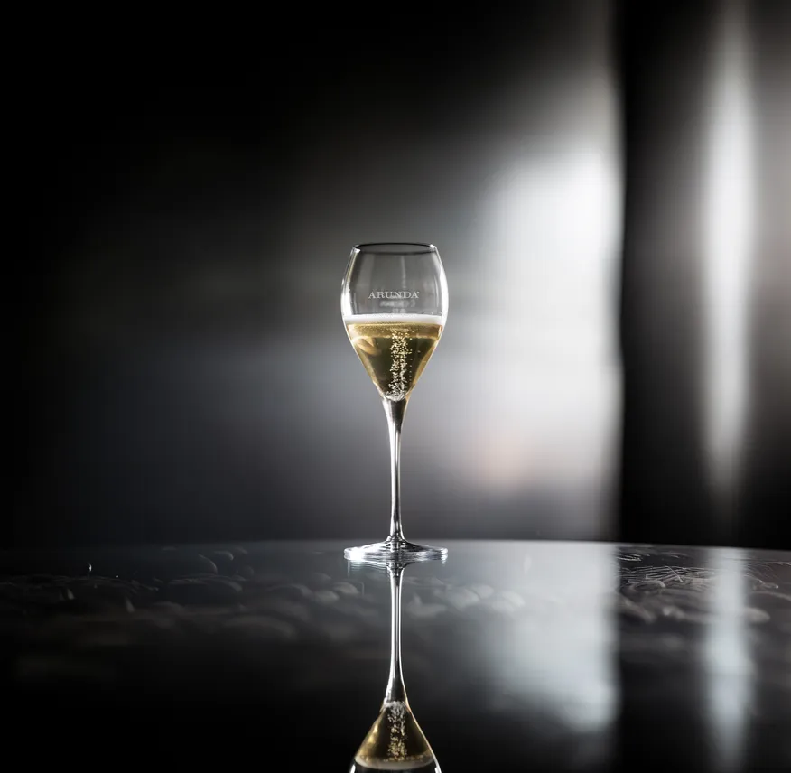 Tulpenförmiges Arunda-Sektglas mit goldenem Sekt und Perlage auf dunkler Spiegelfläche, dramatisches Spotlight
