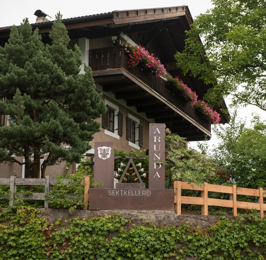 Traditionelles Südtiroler Haus mit Holzbalkonen und rosa Geranien, Arunda Sektkellerei Schild, Mölten