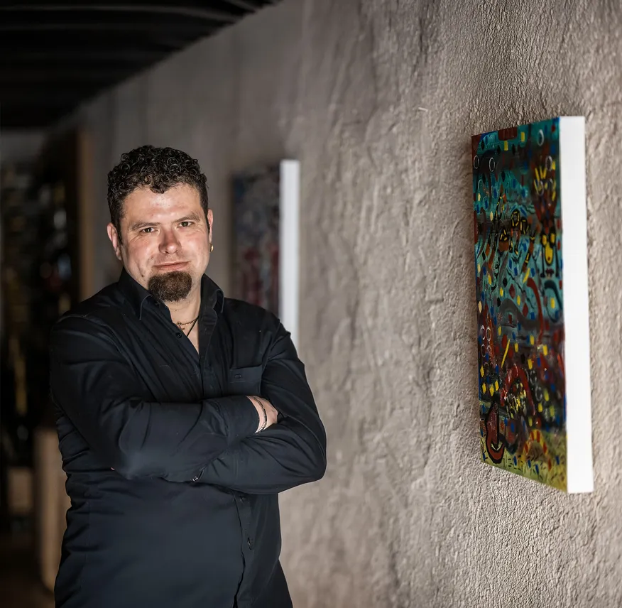 Michael Muggi Reiterer mit verschränkten Armen im Arunda-Kellergang vor farbenfrohem abstrakten Gemälde, dramatisches Licht